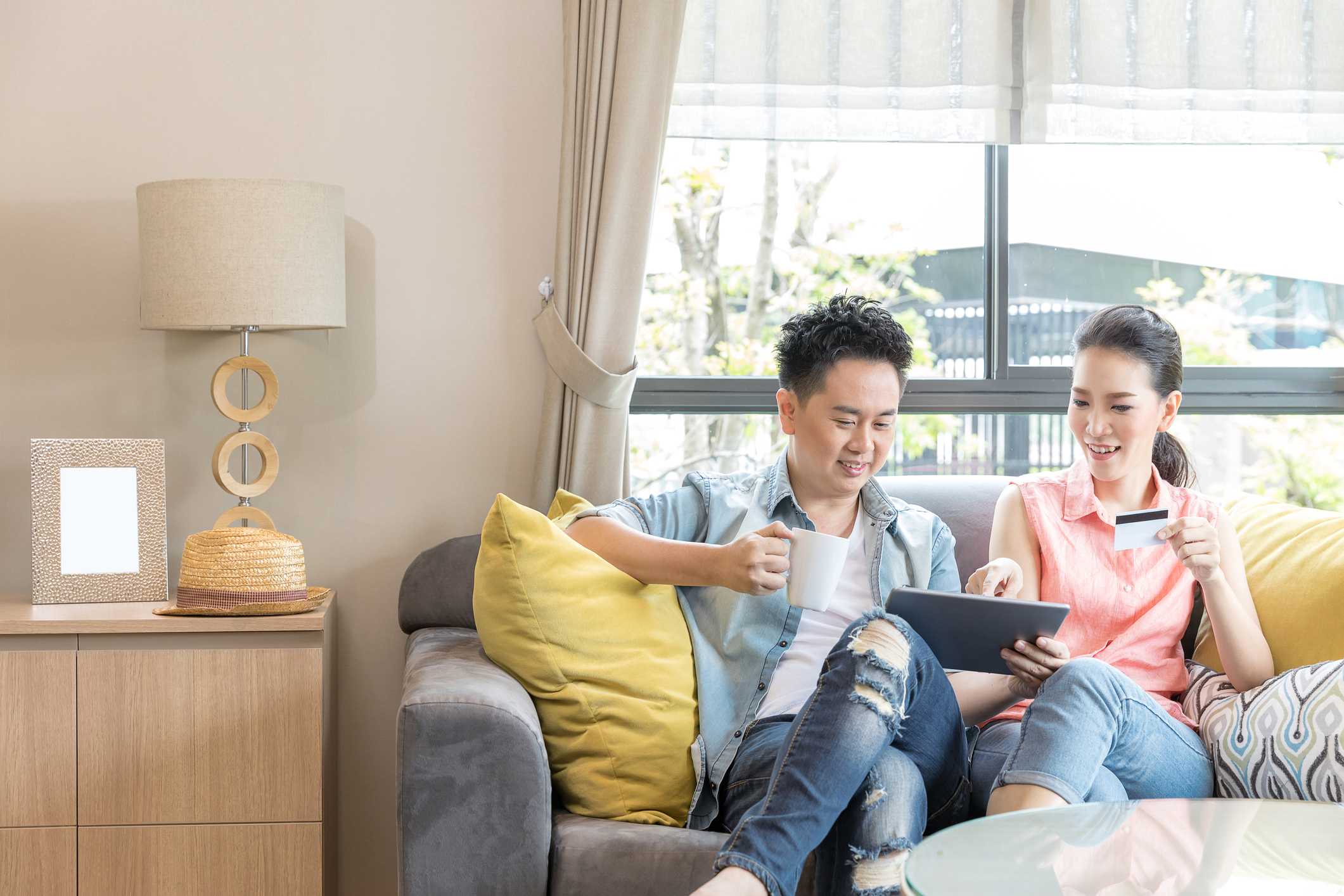 Couple enjoying time together on a couch, using a tablet and a credit card, with a lamp and decorative items in a cozy living room setting.