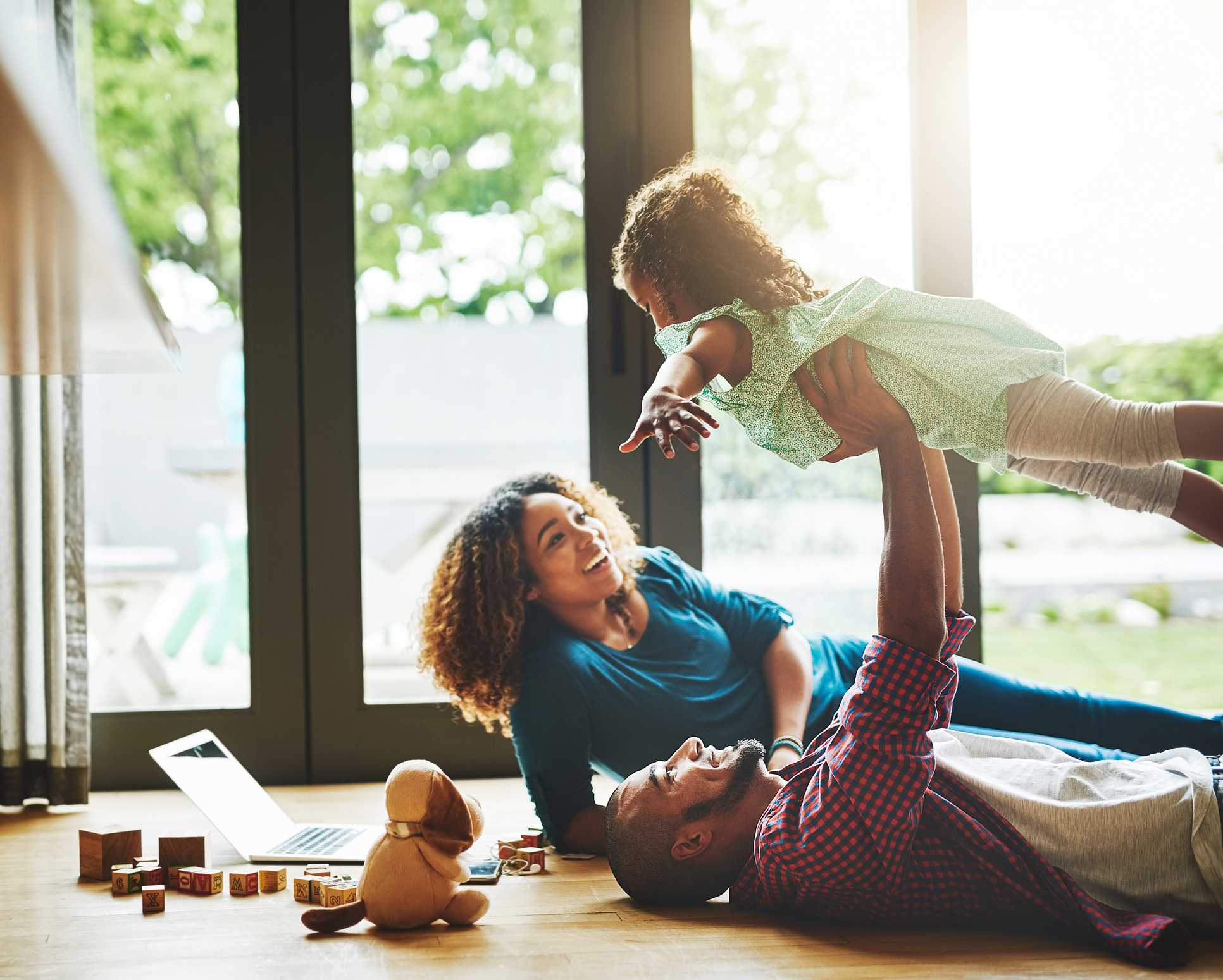 Family enjoying quality time indoors, with a father lifting a young girl playfully, while a mother watches, surrounded by toys and a laptop, highlighting a comfortable home environment.