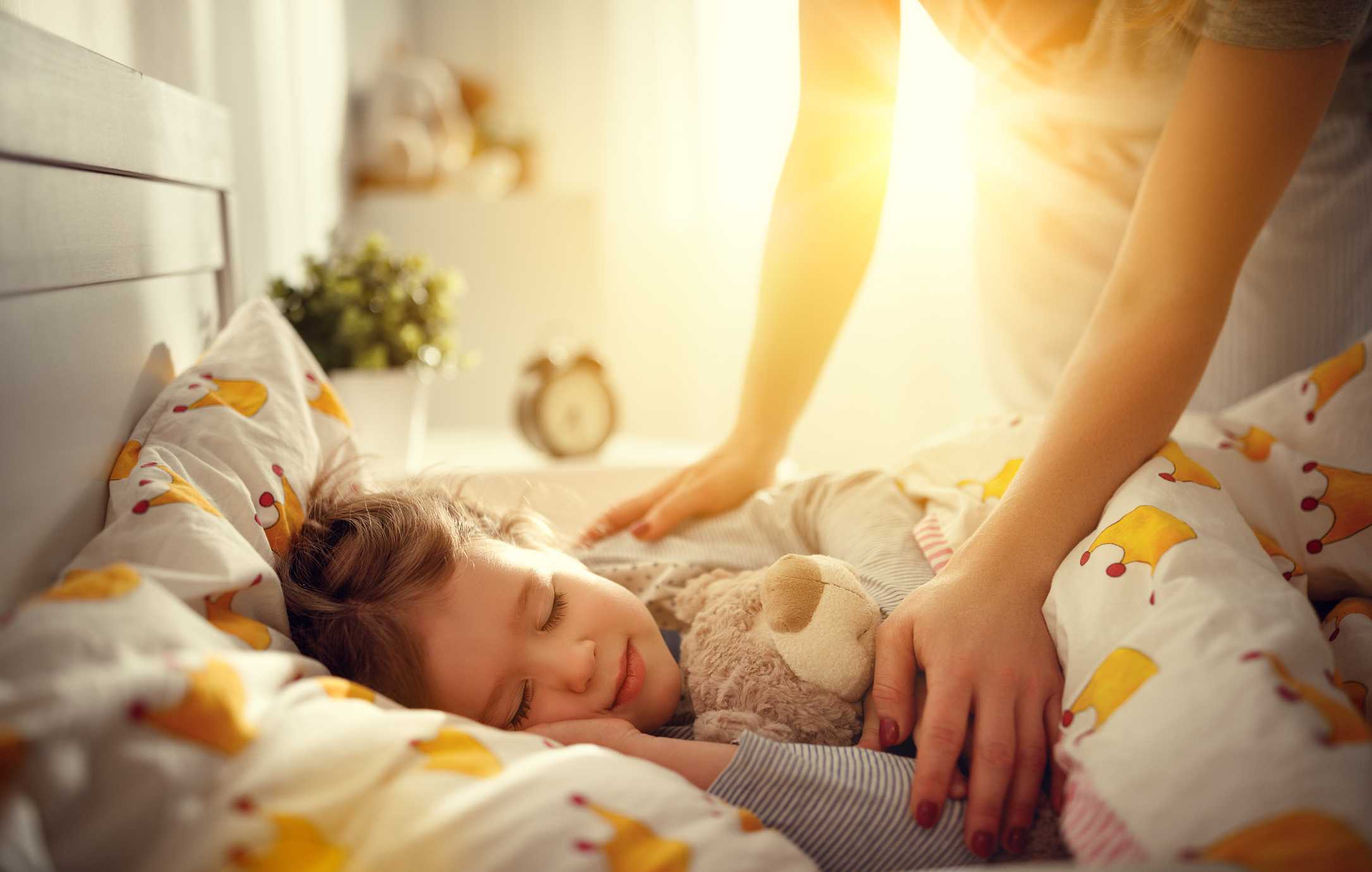 Child sleeping peacefully in bed with a teddy bear, cozy bedding, and a parent gently tucking them in, illustrating the importance of a comfortable home environment for family well-being.